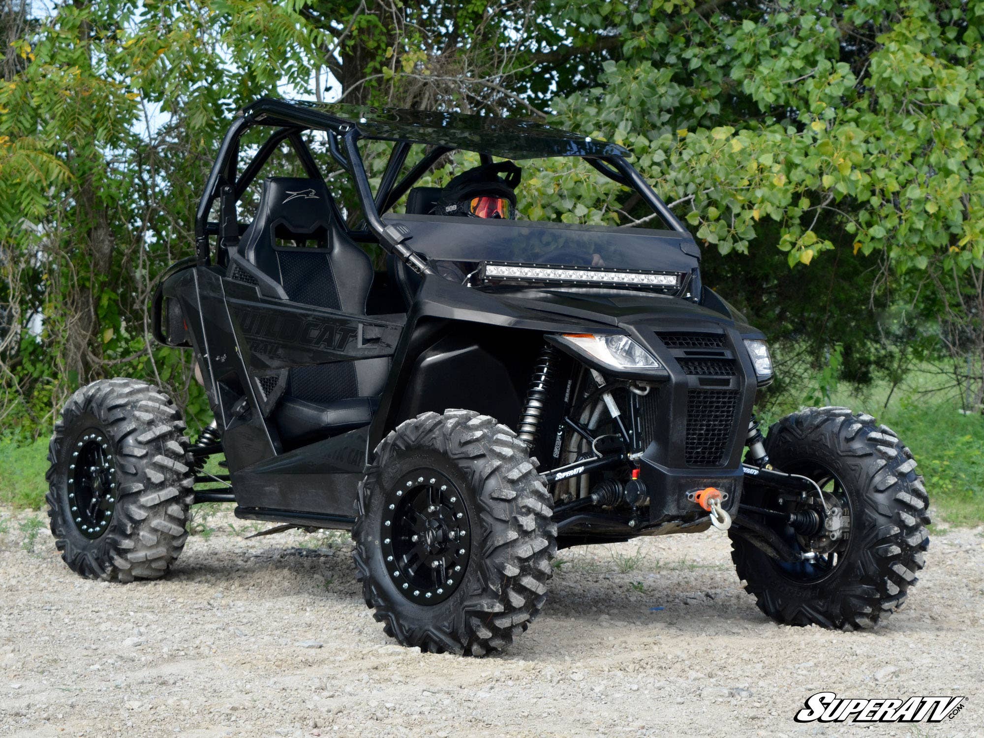An Arctic Cat Wildcat Trail equipped with a 5 Long Travel Kit is parked on gravel. Its black body features angular panels, rugged tires, and a protective roll cage, with dense green foliage in the background. SuperATV logo appears lower right.