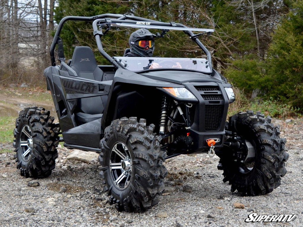 A person wearing a helmet drives a black Arctic Cat Wildcat Trail Sport Flip Windshield side-by-side ATV with aggressive off-road tires over rocky terrain, surrounded by trees and grass, highlighting its rugged, adventurous design.