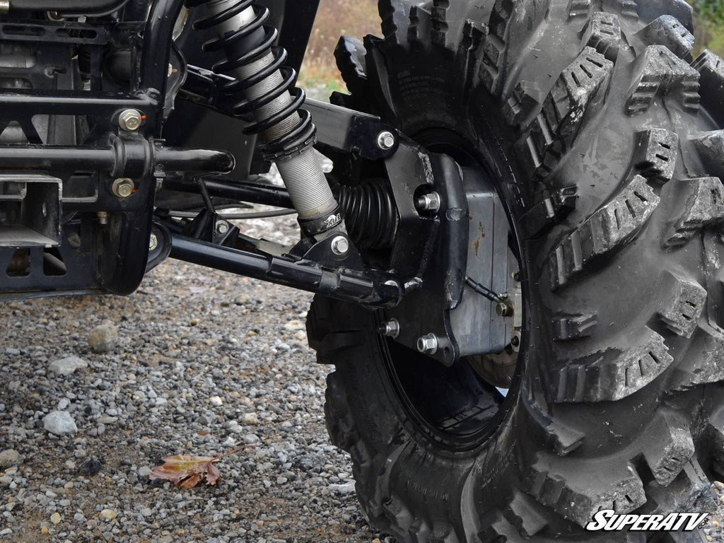 Close-up of an Arctic Cat Wildcat Trail 4 Portal Gear Lift suspension and large, rugged tire with deep treads. Visible are the shock absorber, control arm, and axle on rocky ground. SuperATV logo is in the bottom right corner.