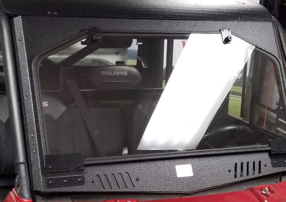 A close-up of the Replacement Windshield Glass Armor Tech on a Polaris off-road vehicle. The glass is mounted in a black frame with visible hinges and latches. Seats and part of the dashboard are visible through the windshield.