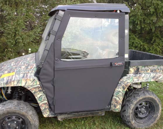 A utility terrain vehicle with a hard roof and Arctic Cat Prowler Convertible Doors featuring clear plastic windows is parked on grass, with evergreen trees in the background. The front wheel and part of the cargo bed are visible.