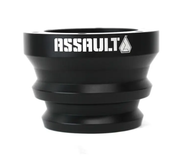 The Assault Industries Steering Wheel Hub is a black, cylindrical metal spacer with beveled edges and grooves. ASSAULT and a geometric logo are printed in white on the top rim. The hub is displayed against a plain white background.