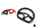 On the right is an Assault Industries Universal Ballistic D V2 UTV Steering Wheel—black, flat-bottomed, with a leather grip and metal center. On the left are two arc-shaped mounting plates (black and red) with holes. A geometric logo appears bottom right.
