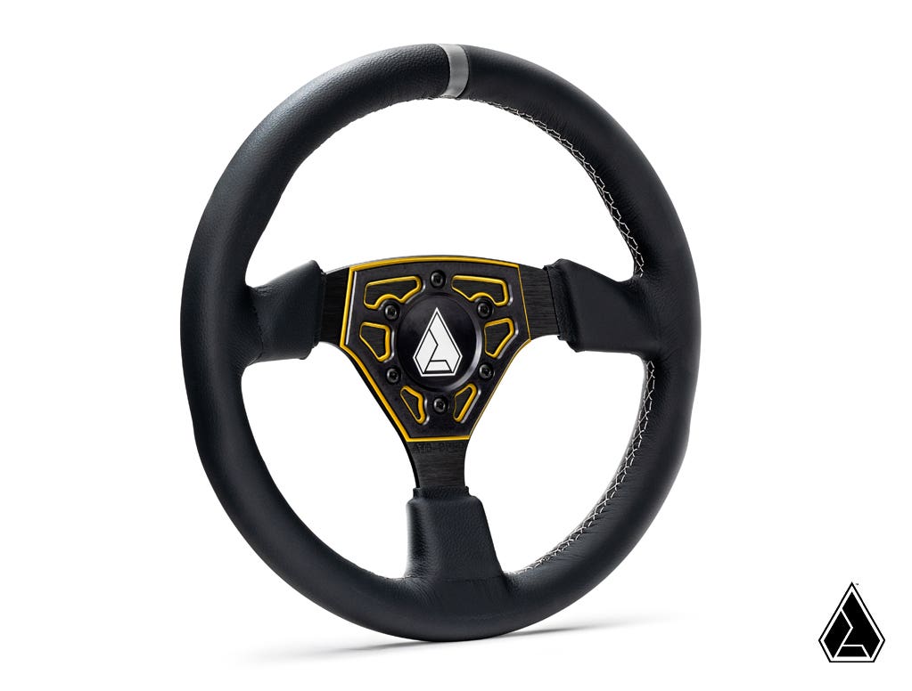 The Assault Industries Universal Navigator Leather UTV Steering Wheel features black leather, a yellow-trimmed three-spoke metal center, decorative stitched grip, and a logo in the middle and the bottom right against a white background.