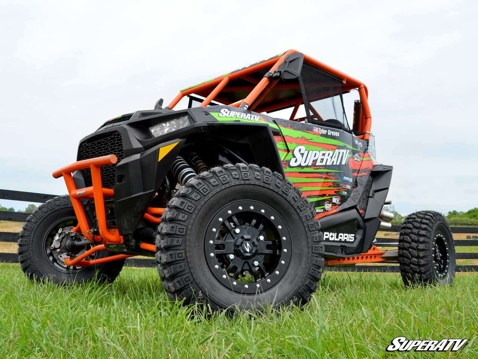 A Polaris off-road vehicle sits on grass, equipped with large SuperATV AT Warrior ATV/UTV Tires. It features a black and orange frame, SuperATV-branded wrap, safety netting, and is set against wooden fences and a cloudy sky.
