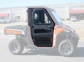 A side view of Full Steel Doors By Armor Tech, installed on a Polaris Ranger 570/500 (Mid-Size) with large off-road tires and a closed black-framed door, parked in a lot with vehicles and commercial buildings in the background.