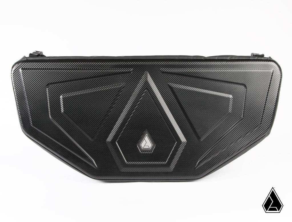 The Assault Industries RUXAK Deck Pak for Recon Rack (Can-Am Maverick X3) is a black, textured hard-shell case with an angular geometric pattern and a small triangular logo centered on the front. The background is white with a matching logo at the bottom 