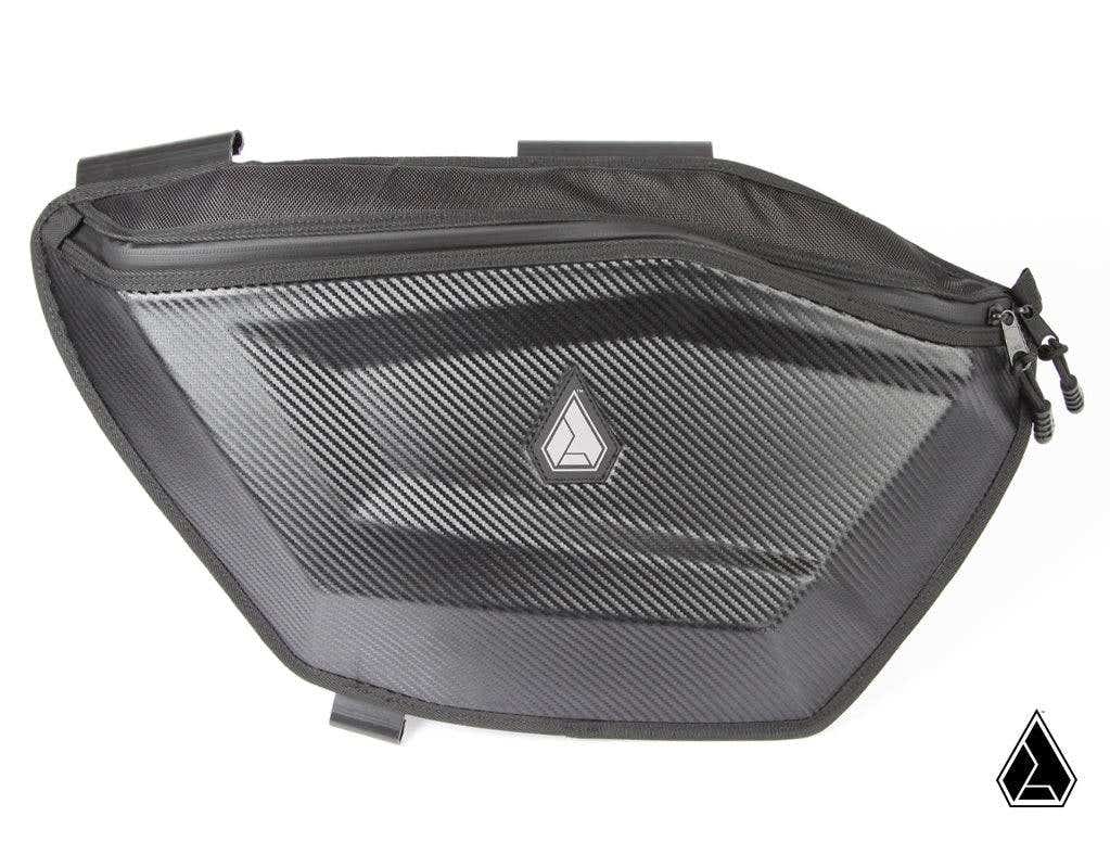 The Assault Industries Polaris RZR Pro R RUXAK Door Paks feature a hard-shell, black design with carbon fiber texture, a small geometric logo, reinforced corners, top zipper, and secure attachment points for your vehicle.