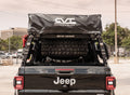 Rear view of a black Jeep with a spare tire, rooftop tent in a black cover, and ROAD ARMOR text on the adjustable Treck 5Ft. Mid-Size Truck Bed Rack System (Bracket Kit Not Included) in texture black, outdoors by a building and fence.