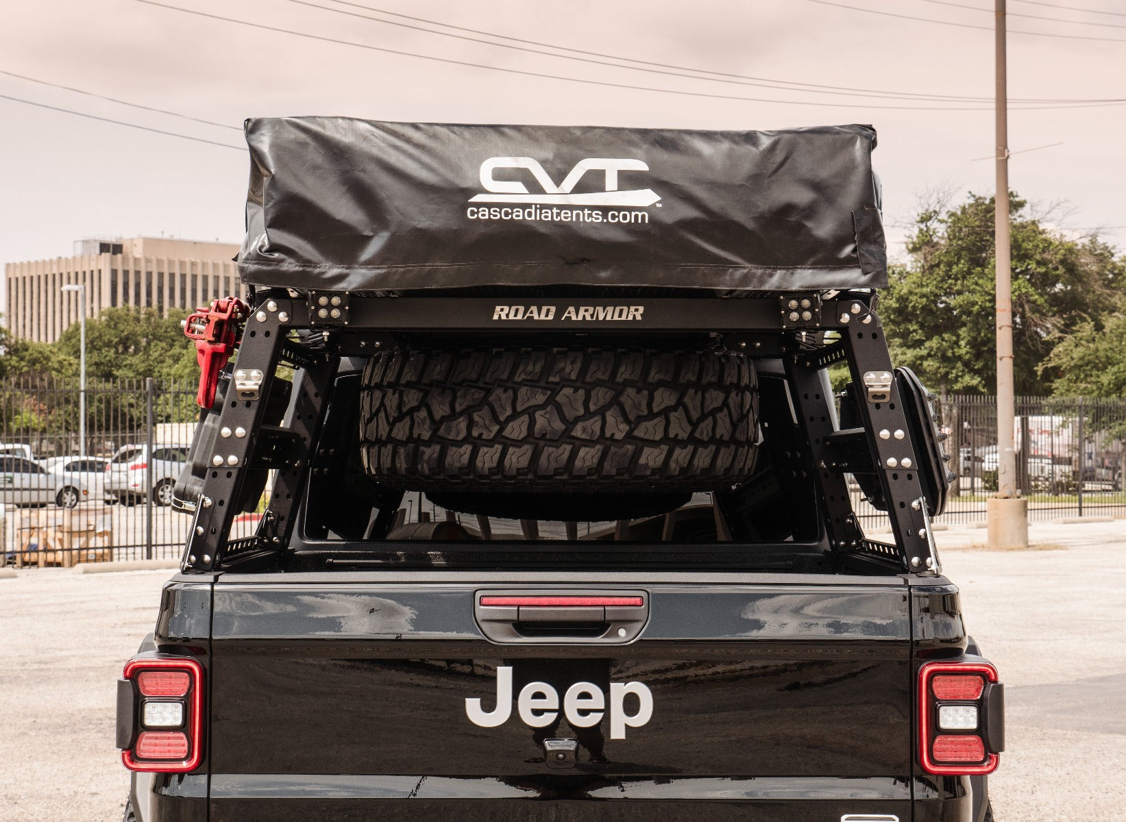 Rear view of a black Jeep with a spare tire, rooftop tent in a black cover, and ROAD ARMOR text on the adjustable Treck 5Ft. Mid-Size Truck Bed Rack System (Bracket Kit Not Included) in texture black, outdoors by a building and fence.