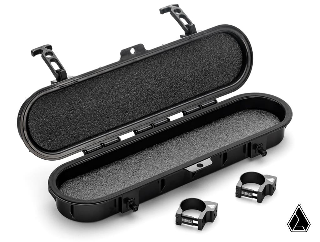 The Assault Industries UTV Belt Case is a black, hard-shell oval case with foam padding, two latches, a textured surface, and a geometric triangular logo in the bottom right corner. Two metal mounting brackets are displayed in front of the open case.