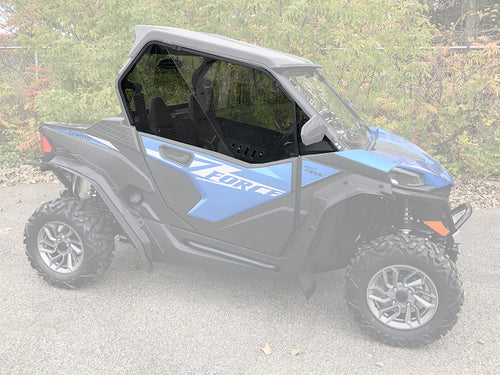 A CF Moto Z-Force 800-950 side-by-side with rugged tires and sporty graphics is parked on pavement, featuring the 2023+ Trail Vented Scratch Resistant Polycarbonate Upper Doors Kit (Tinted); greenery and a chain-link fence are in the background.