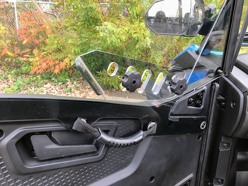 Close-up of a CF Moto Z-Force 800-950 Trail Vented Scratch Resistant Polycarbonate Upper Doors Kit (Tinted, 2023+) with black interior, clear half window secured by knob fasteners, handle, strap, and textured panel. Green and orange foliage outside.