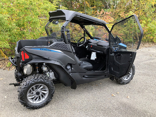 A black CF Moto Z-Force 800-950 Trail (2023+) features tinted, vented, scratch-resistant polycarbonate upper doors kit, with the right door open. Rugged tires and a sporty design stand out against lush greenery and autumn-colored trees.