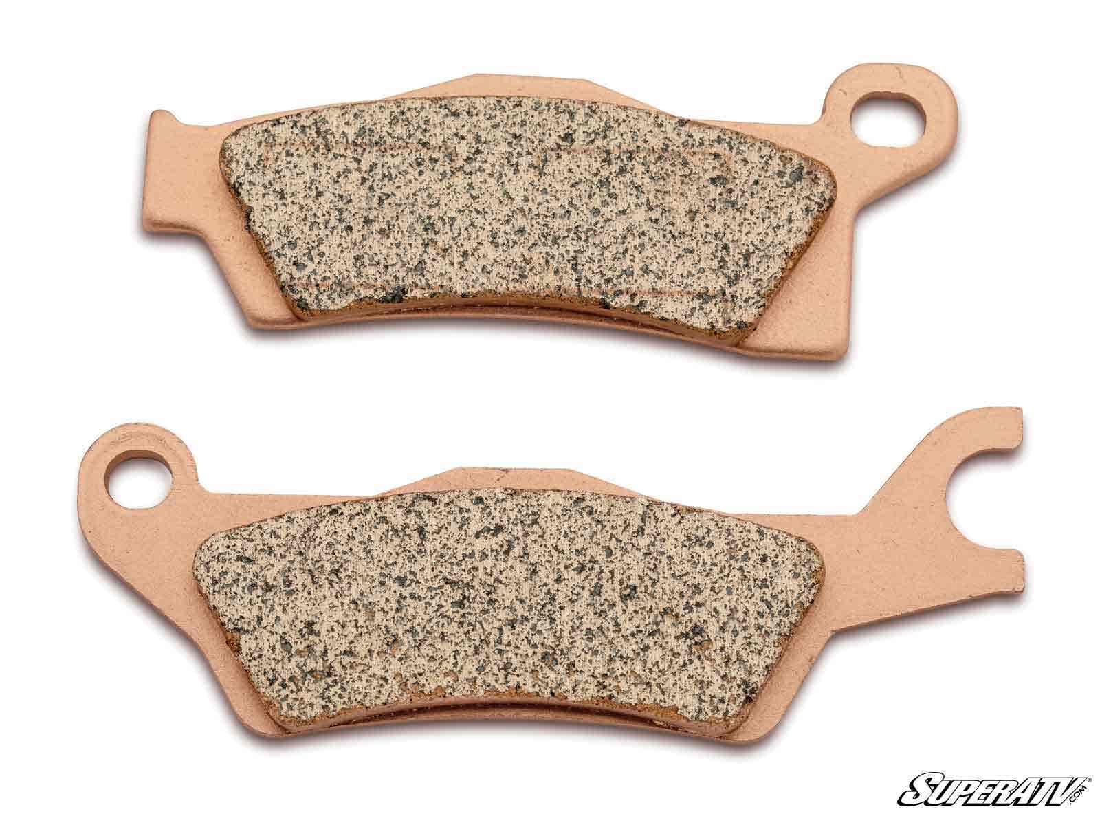 Two Can-Am Outlander Sintered Brake Pads with textured friction surfaces are shown side by side on a white background. Each pad features unique shapes with notches and holes for mounting. The SuperATV logo is in the bottom right corner.
