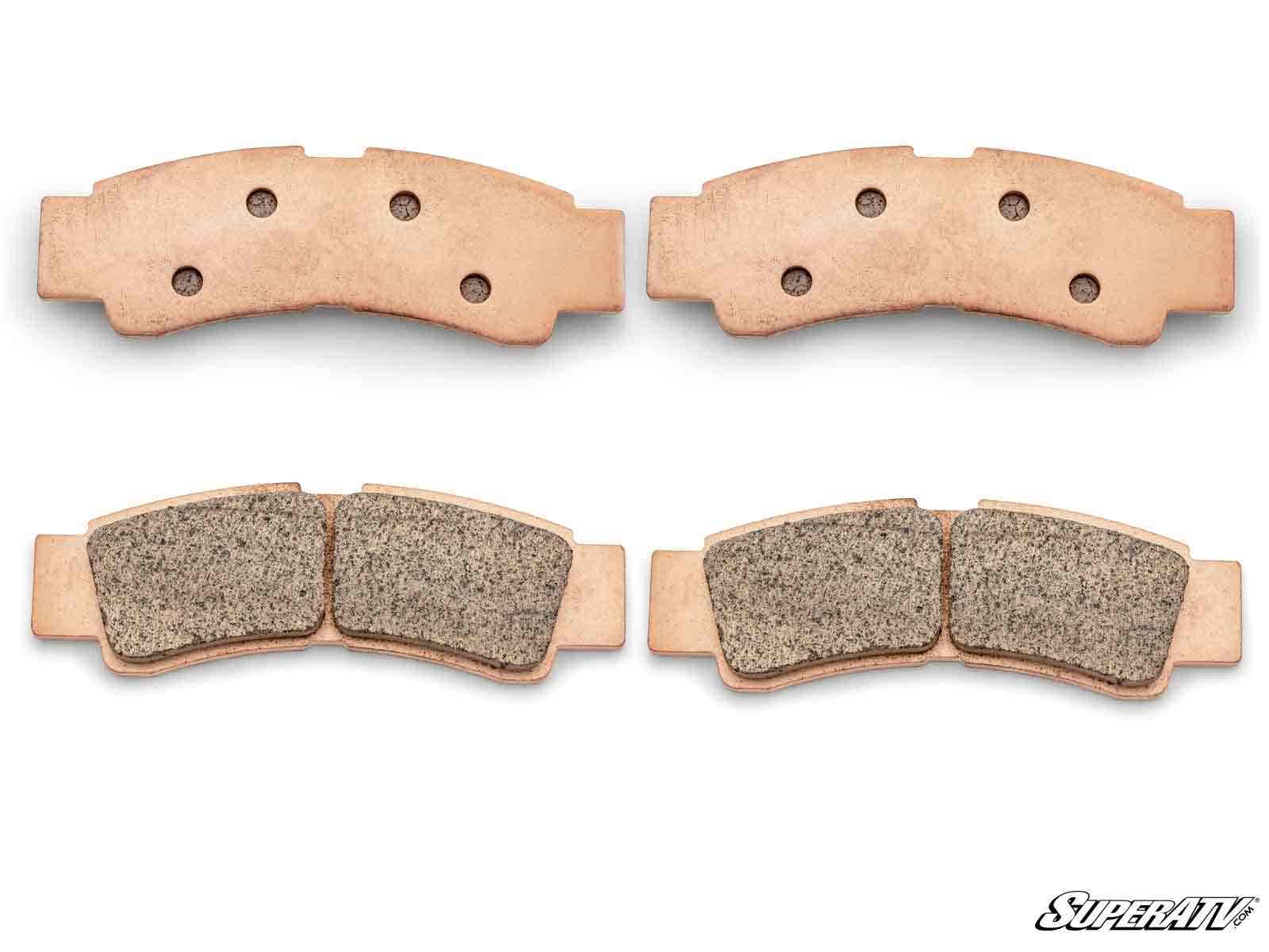 Four Kawasaki Teryx KRX Sintered Brake Pads are arranged in two pairs on a white background. Each pad features a curved shape, visible holes on the back, and textured friction material. The “SuperATV” logo appears in the bottom right corner.