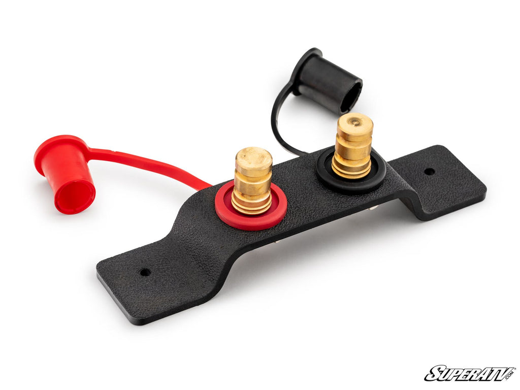 The Battery Terminals Relocation Kit includes a black rectangular mounting bracket with two brass terminals: one with a red ring and cap (positive) and one with a black ring and cap (negative), each with wire-attached covers. SuperATV logo at lower right.