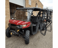 A Can Am Defender Max Full Hard Cab Enclosure in red and black with large tires is parked on pavement near a brick building. The front passenger and rear doors are open, showing multiple seats. The windshield is clear, and a winch is mounted on the front.