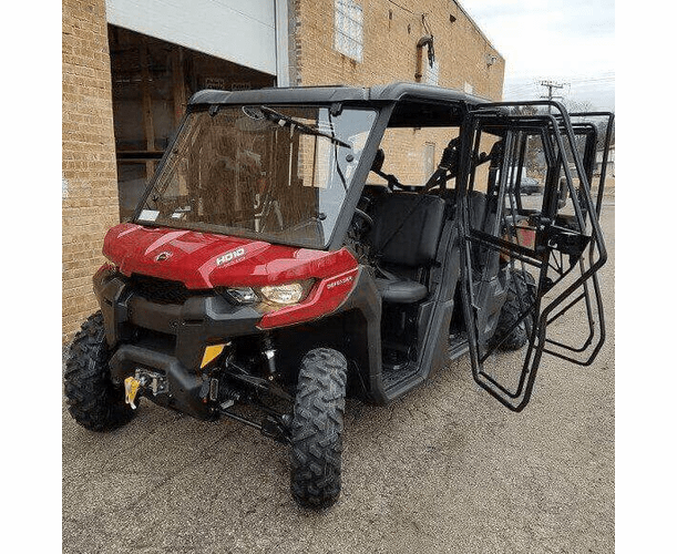 A Can Am Defender Max Full Hard Cab Enclosure in red and black with large tires is parked on pavement near a brick building. The front passenger and rear doors are open, showing multiple seats. The windshield is clear, and a winch is mounted on the front.