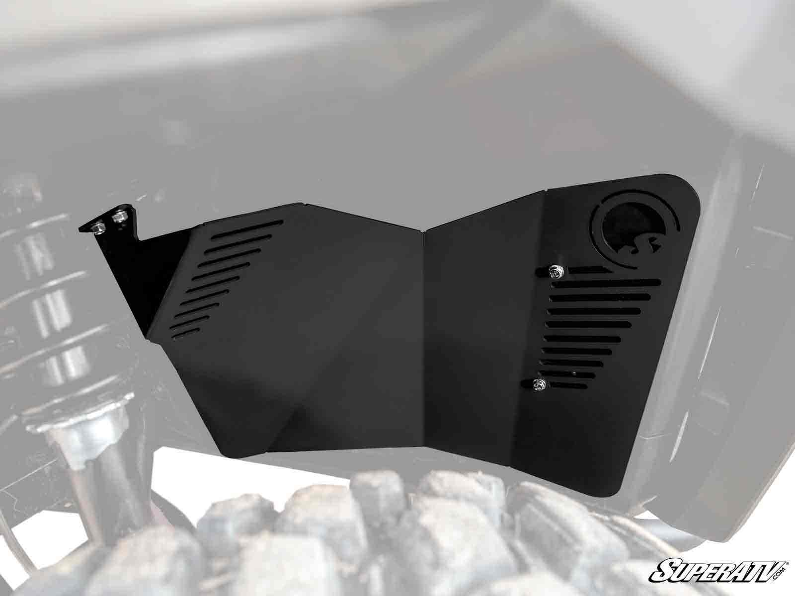 A black Can-Am Defender HD8 Inner Fender Guard with vent slots and a circular cap opening is mounted under a vehicle near the suspension and tire. The background is partially faded, with the SuperATV logo in the lower right corner.