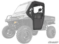 A Can-Am Defender Primal Soft Cab Enclosure Door in black with a clear window is installed on the driver side of a rugged, lifted off-road utility vehicle; the cab area is enclosed, and the rest of the vehicle appears semi-transparent.