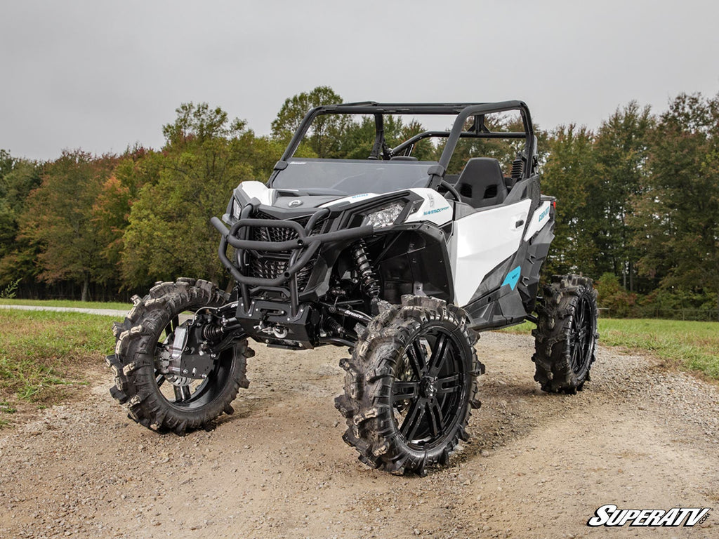 A Can-Am Maverick Sport 6 Portal Gear Lift with a rugged white and black exterior, roll cage, and large mud tires is parked on a gravel path, surrounded by green grass and dense trees beneath a cloudy sky.