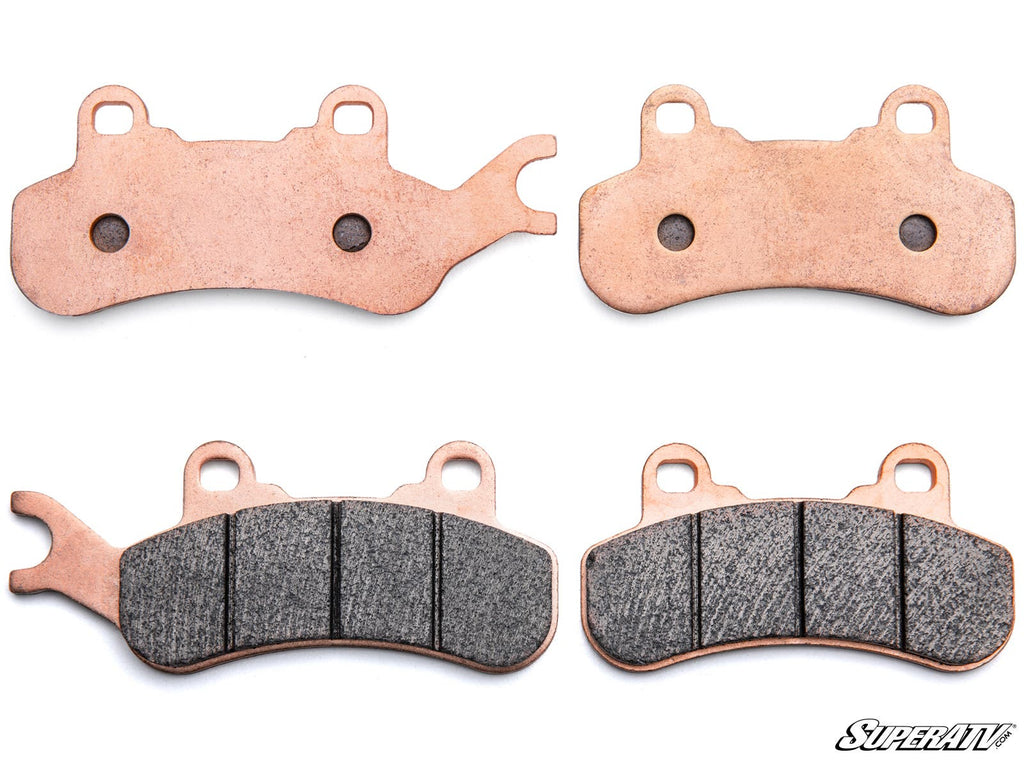 Four Can-Am Maverick X3 Sintered Brake Pads are shown on a white background. The upper pads display metal backing; the bottom ones show dark friction surfaces, with an extra tab on the bottom left. SuperATV appears in the lower right corner.