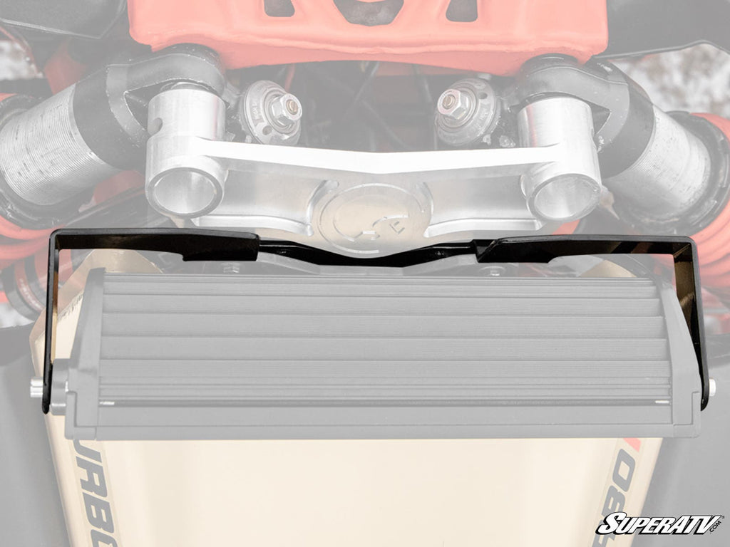 A close-up of a vehicles front suspension and steering with a semi-transparent outline of the Can-Am Maverick X3 12 Shock Tower Light Bar Mount; metallic arms, bolts, and SUPERATV branding appear in the bottom right corner.