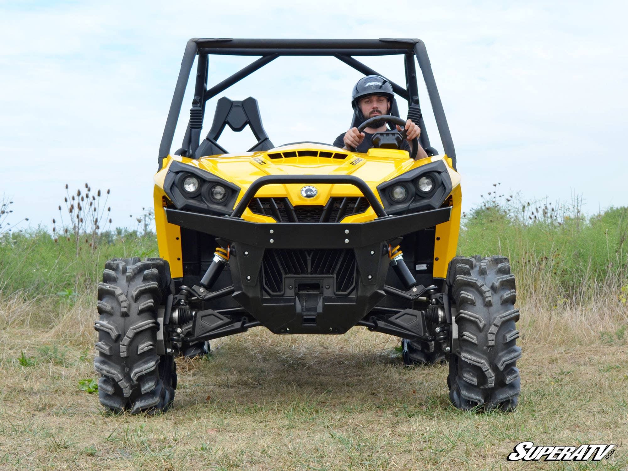 A helmeted driver operates a bright yellow off-road vehicle with rugged tires on grass under a partly cloudy sky, showcasing the Can-Am Commander 4 Portal Gear Lift. The SuperATV logo appears in the bottom right corner.