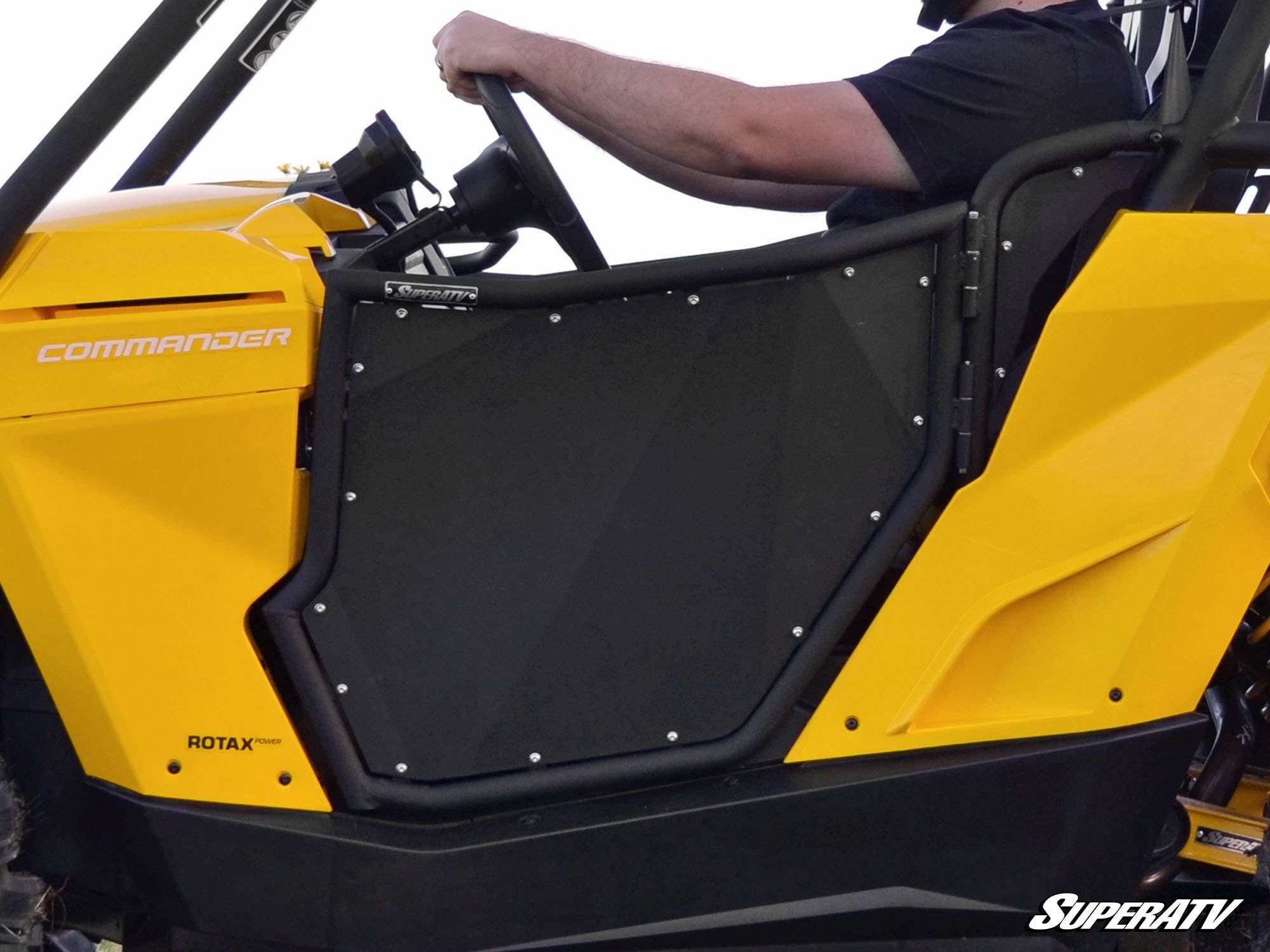 A person in a black shirt drives a bright yellow off-road vehicle equipped with Can-Am Commander Aluminum Doors. The vehicle features the SuperATV logo at the bottom right, with part of the steering wheel and dashboard visible.