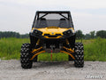 A yellow off-road ATV with large, rugged tires and black accents is parked on gravel under an overcast sky. It features the Can-Am Commander Half Windshield. Green grass and trees are in the background, with the SuperATV logo in the lower right corner.