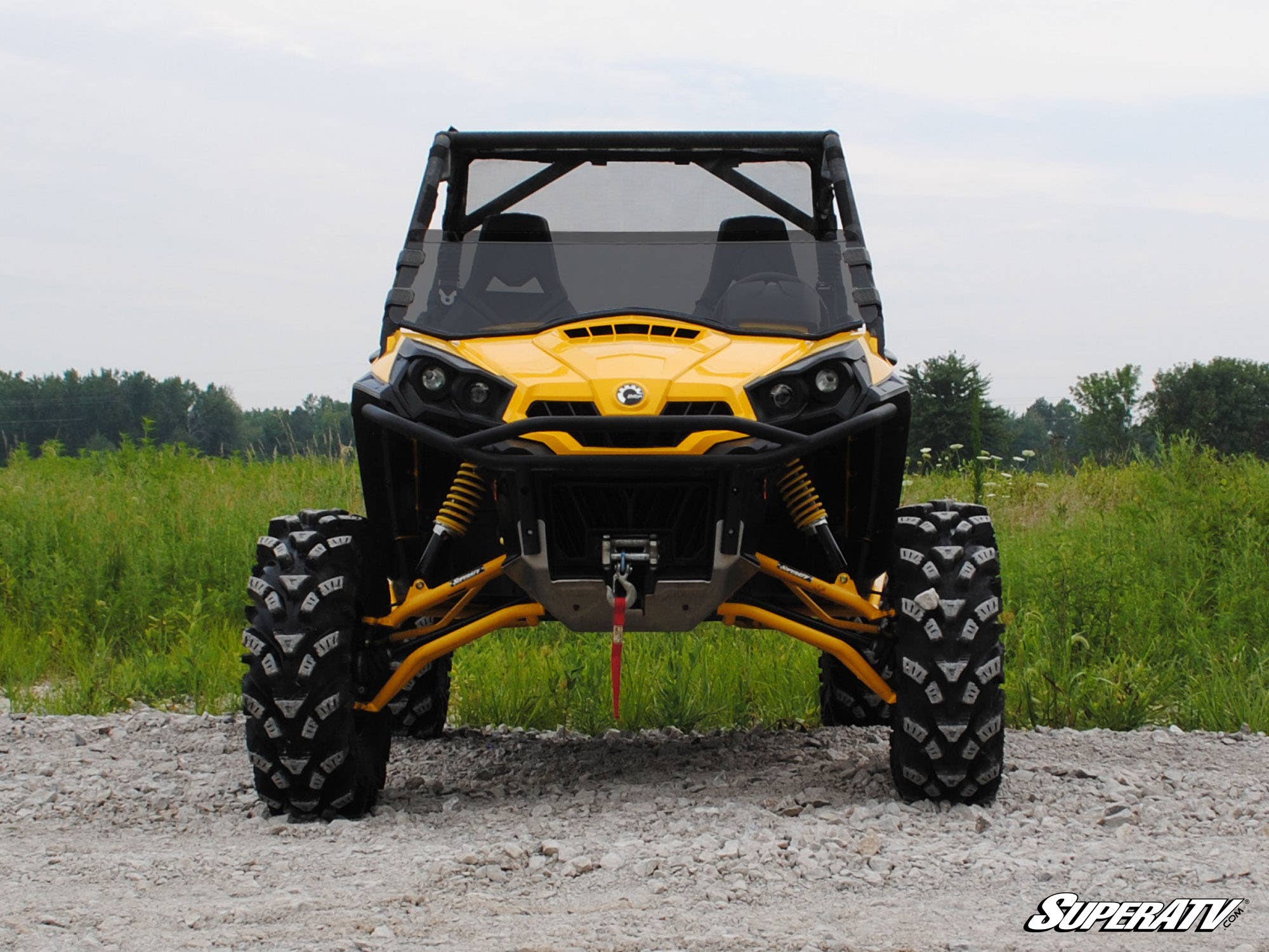 A yellow off-road ATV with large, rugged tires and black accents is parked on gravel under an overcast sky. It features the Can-Am Commander Half Windshield. Green grass and trees are in the background, with the SuperATV logo in the lower right corner.