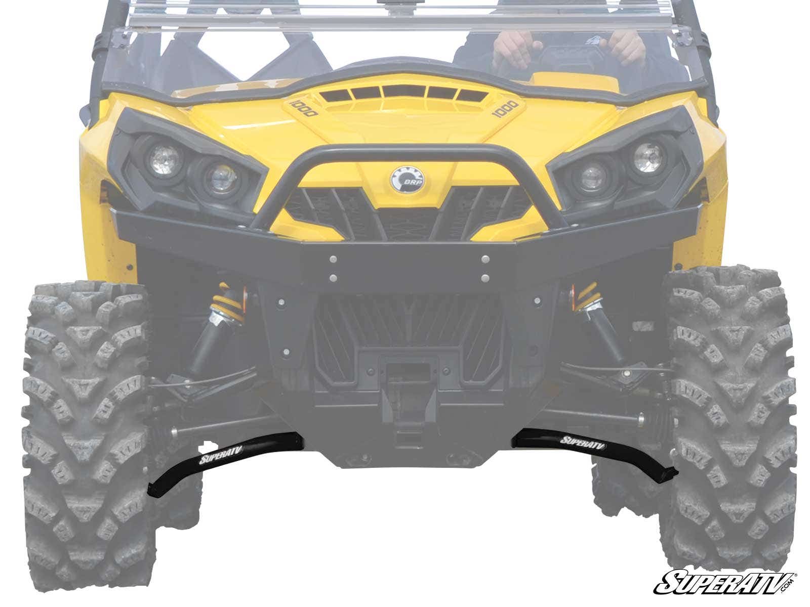 Close-up front view of a yellow off-road utility vehicle showing rugged tires and Can-Am Commander High Clearance Lower A-Arms by SuperATV. The black suspension components are highlighted, with the SuperATV logo at the bottom right corner.