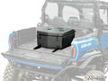 A Can-Am Commander Cooler/Cargo Box is mounted in the bed of a blue Can-Am off-road vehicle, secured with metal latches and brackets. The tailgate is down, and the SuperATV logo appears in the bottom right corner.