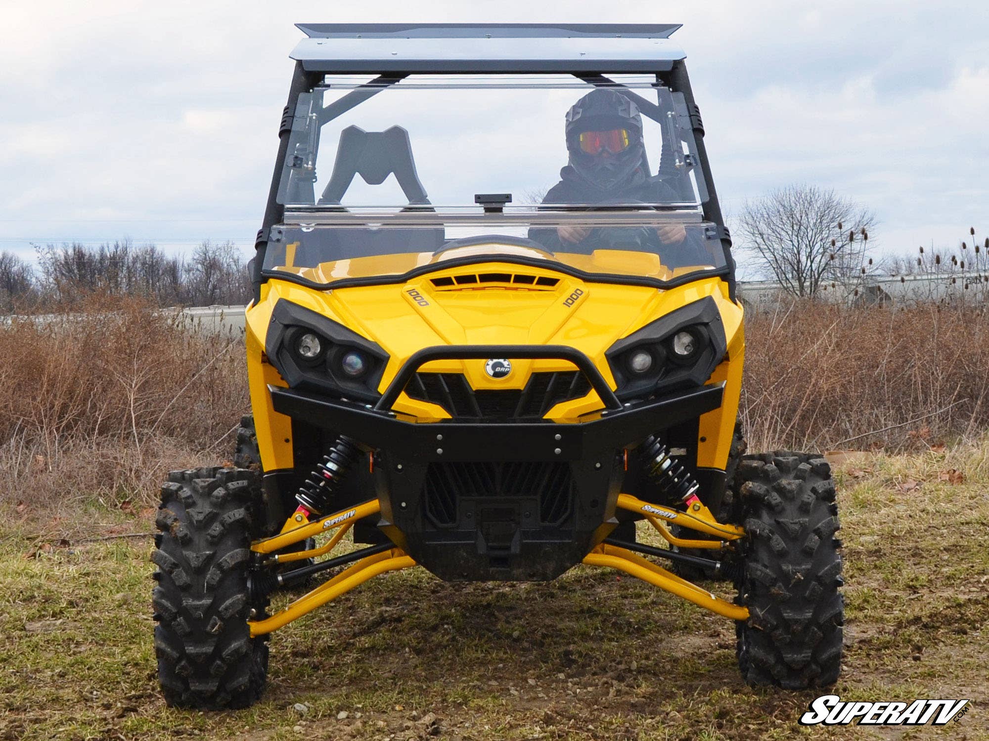 A yellow off-road side-by-side ATV with large tires is parked on grass. The driver, in helmet and goggles, sits behind a Can-Am Commander Flip Windshield. Brown shrubs and a cloudy sky are in the background. The SuperATV logo is bottom right.