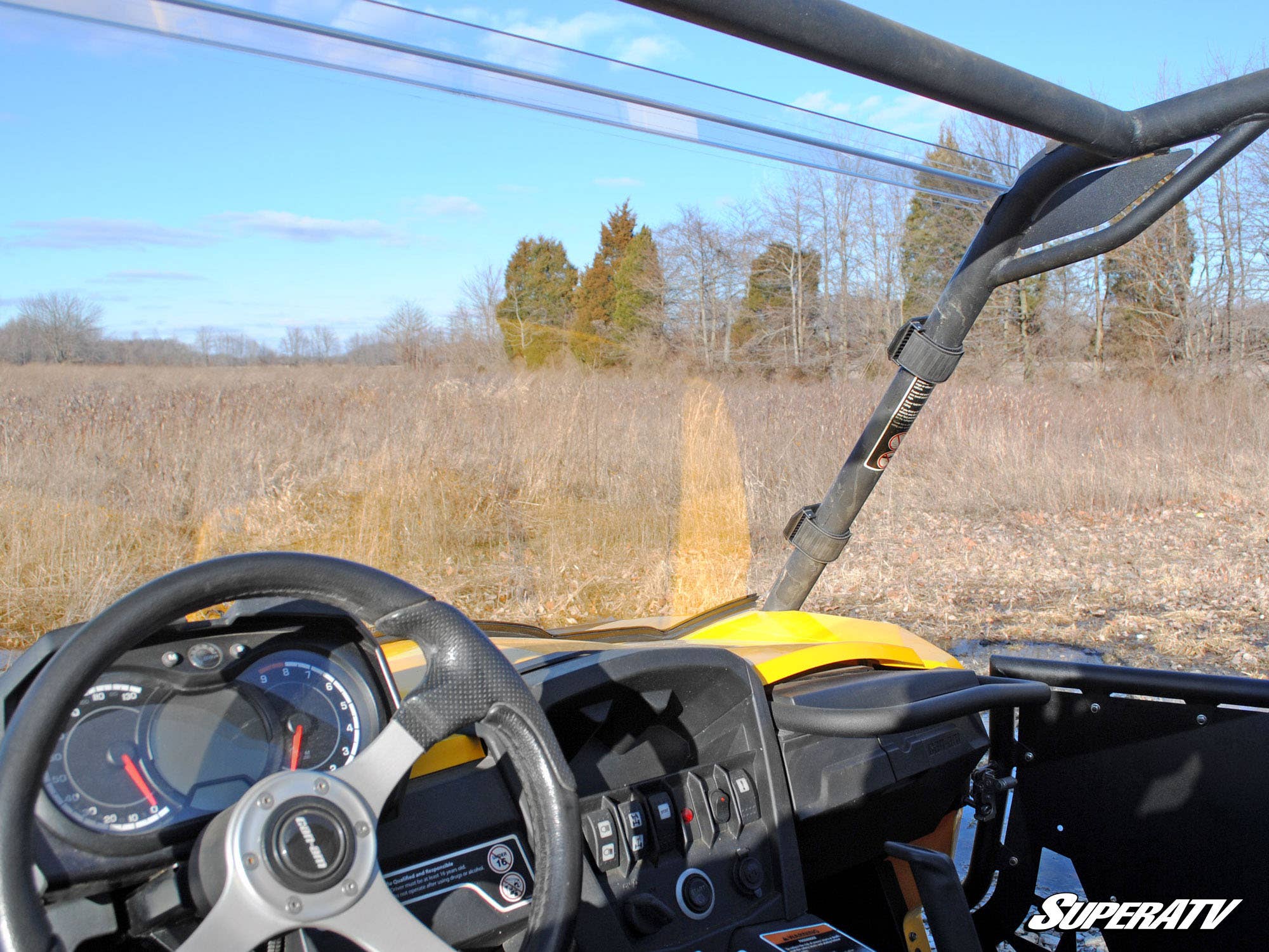 View from inside a yellow off-road vehicle featuring the Can-Am Commander Full Windshield, steering wheel, dashboard, and roll cage. The split windshield frames a dry, grassy field with scattered trees under a clear blue sky. SuperATV logo at bottom right