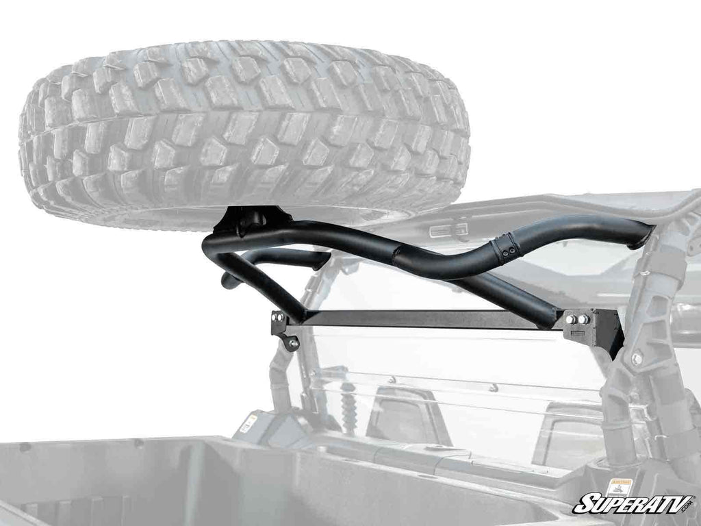 A Can-Am Commander Spare Tire Carrier, made of black metal, is mounted on the rear roll cage of an off-road vehicle, securely holding a large spare tire above the cargo bed. The SuperATV logo appears in the lower right corner.