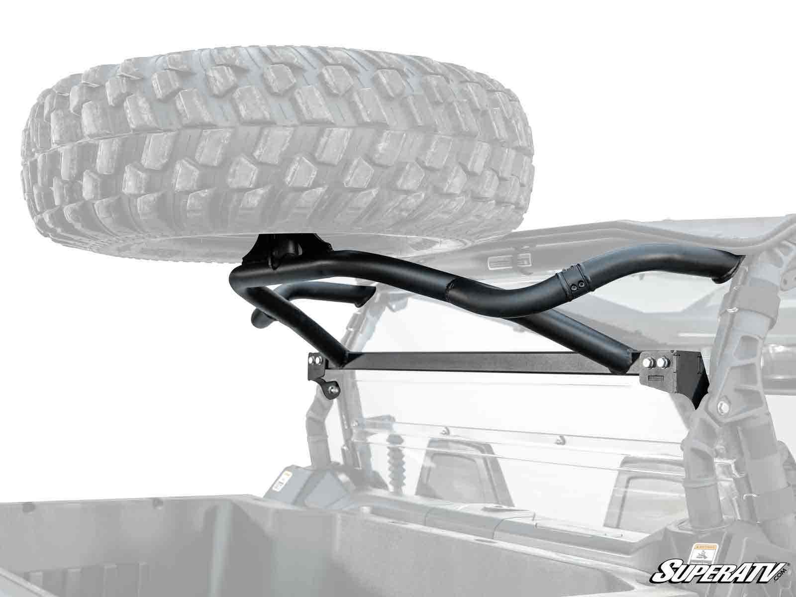 A Can-Am Commander Spare Tire Carrier, made of black metal, is mounted on the rear roll cage of an off-road vehicle, securely holding a large spare tire above the cargo bed. The SuperATV logo appears in the lower right corner.
