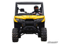 A helmeted driver with goggles sits in a bright yellow off-road utility vehicle, equipped with large tires and black accents. Seen from the front on a white background, the “SuperATV” logo is at the lower right. Product: Can-Am Defender HD10 3 Lift Kit.