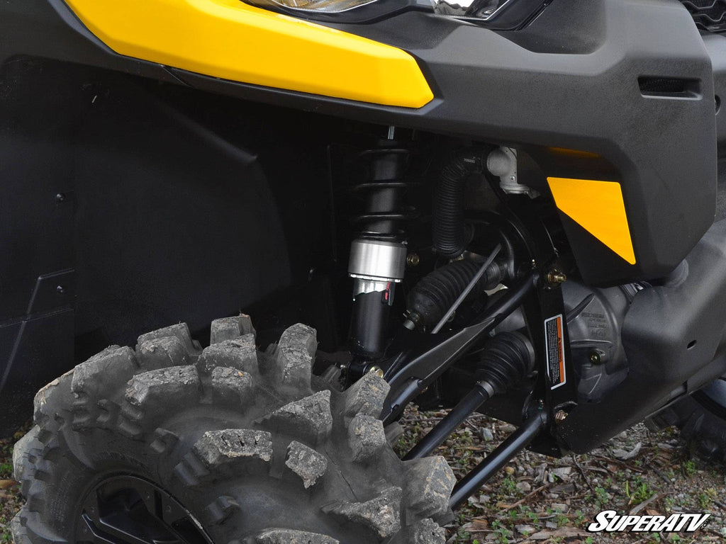 Close-up of a Can-Am Defender HD5 with a 3 Lift Kit, showing the front left wheel’s rugged tire, visible coil spring suspension, and partial yellow-black body panels and headlight. SuperATV logo appears in the bottom right corner.