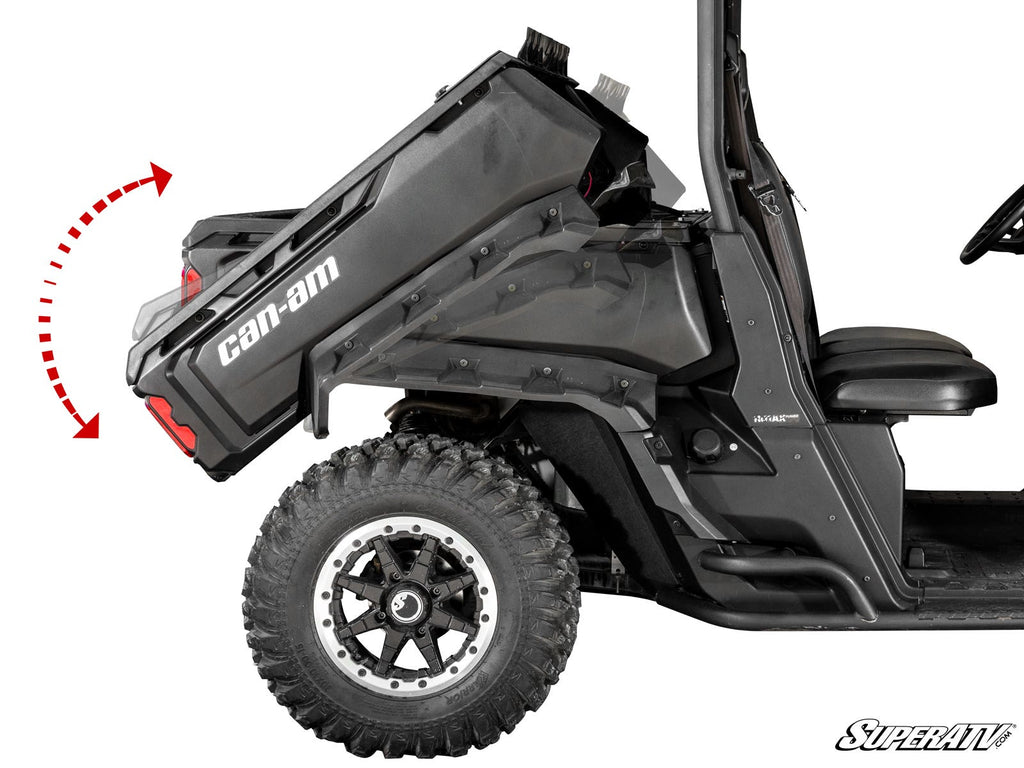 A side view of a Can-Am Defender equipped with the Electric Dump Bed Kit shows its black cargo bed tilted upward, demonstrating the dumping function. A red dashed arrow marks the tilt direction. The vehicle features large rugged tires and visible seating.
