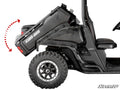 A side view of a Can-Am Defender equipped with the Electric Dump Bed Kit shows its black cargo bed tilted upward, demonstrating the dumping function. A red dashed arrow marks the tilt direction. The vehicle features large rugged tires and visible seating.