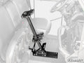 The Can-Am Defender Floor-Mounted Gun Holder is a black metal rack between the seats of a utility vehicle. It holds two guns upright, with nearby seats, steering wheel, and dashboard faintly visible in grayscale.