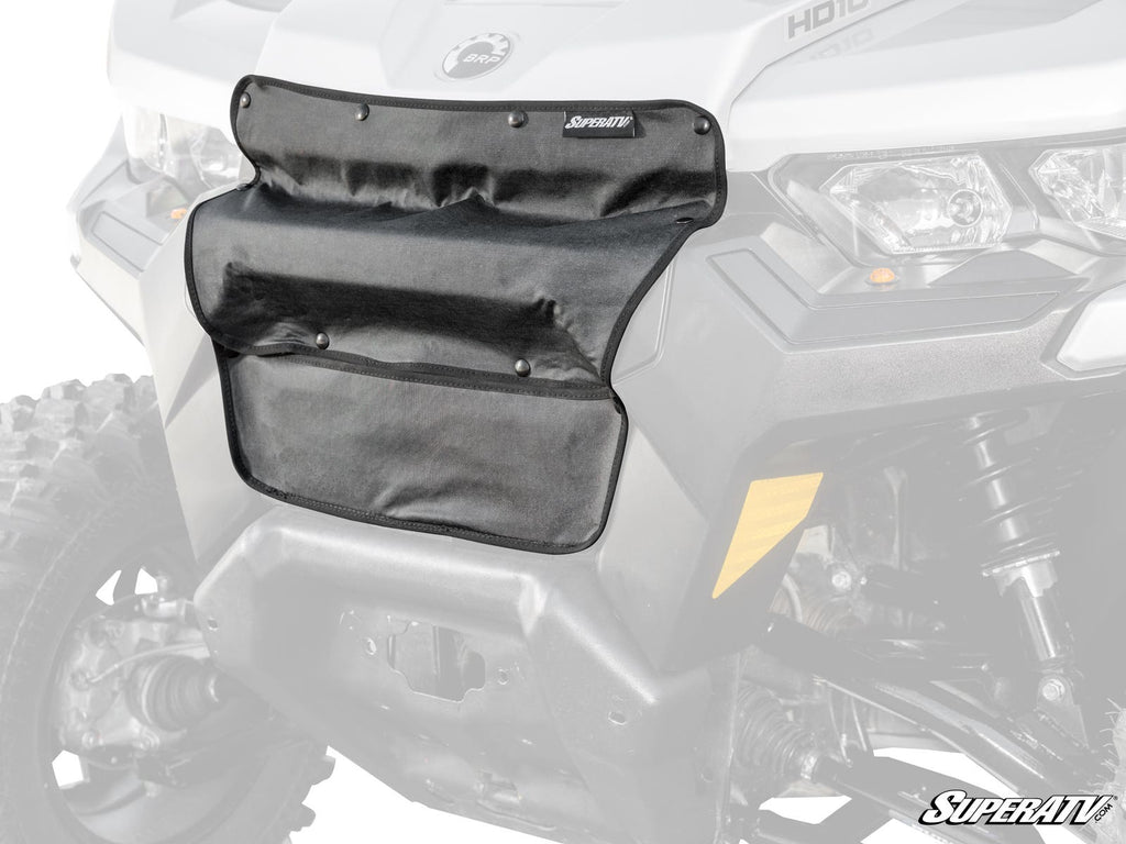 A black Can-Am Defender Grille Cover with multiple pockets and snap closures is attached to the front grill of a gray off-road vehicle, covering the center area. The SuperATV logo is visible in the bottom right corner.