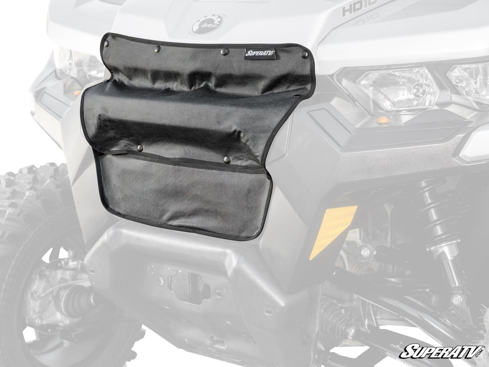 A black Can-Am Defender Grille Cover with multiple pockets and snap closures is attached to the front grill of a gray off-road vehicle, covering the center area. The SuperATV logo is visible in the bottom right corner.