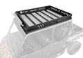 A close-up of the Can-Am Defender MAX Outfitter Roof Rack, a black metal rack with slotted sides and reinforced crossbars, mounted on a camouflage-patterned off-road vehicle against a partially faded background.