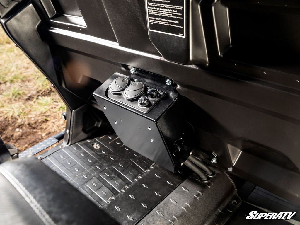 A Can-Am Defender Cab Heater is mounted on the floor behind the front seats. It has two round control dials, two air vents, black textured floor mats nearby, molded black panels, and displays the SuperATV logo.