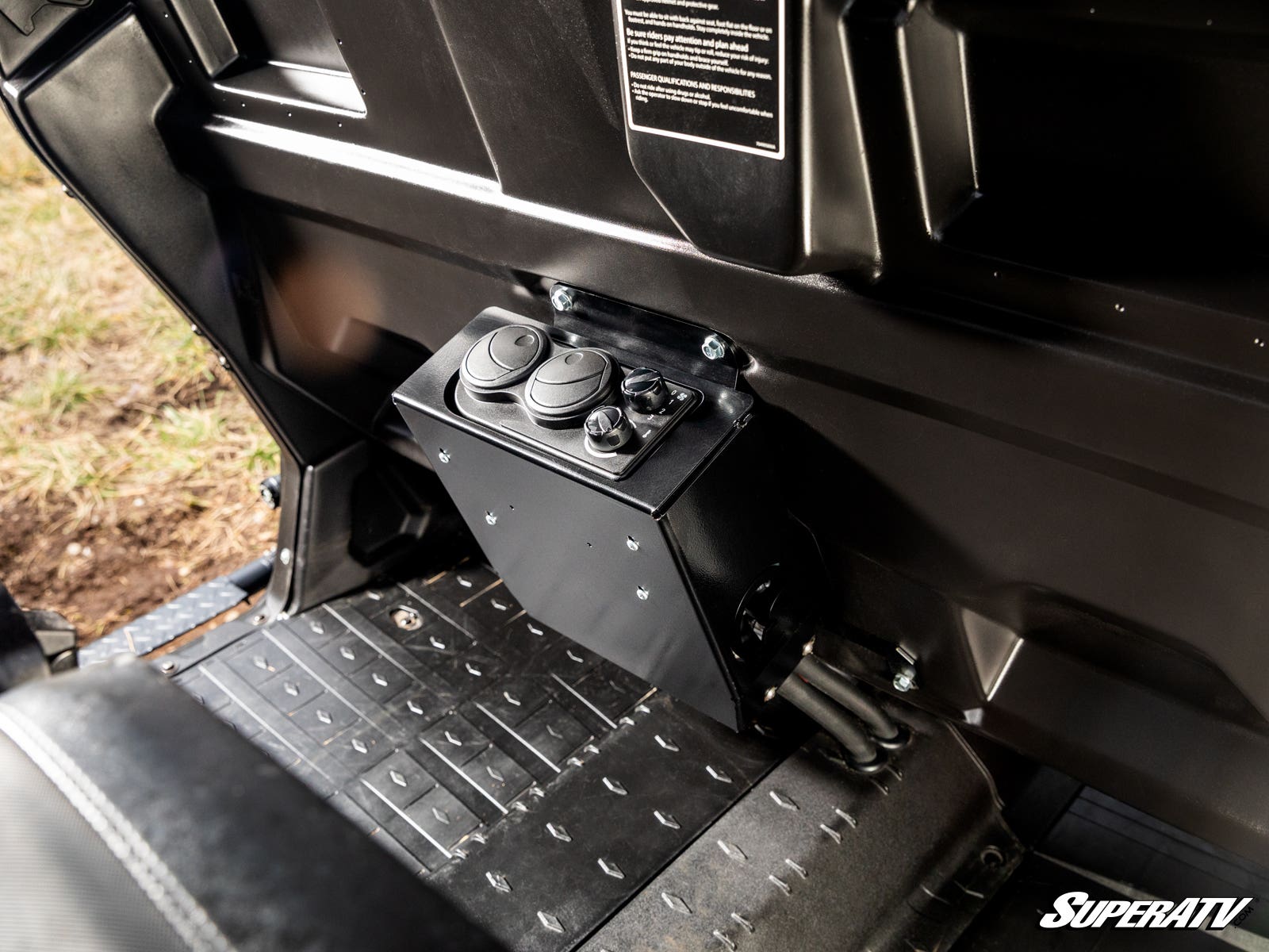 A Can-Am Defender Cab Heater is mounted on the floor behind the front seats. It has two round control dials, two air vents, black textured floor mats nearby, molded black panels, and displays the SuperATV logo.