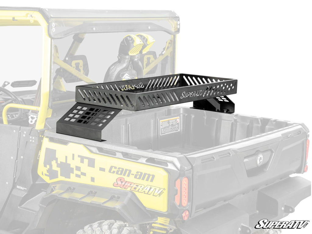 A Can-Am Defender Bed Rack Delta by SuperATV is mounted in the bed of a yellow Can-Am utility vehicle, with the rack elevated above the bed. The SuperATV logo is visible on both the rack and the vehicle.