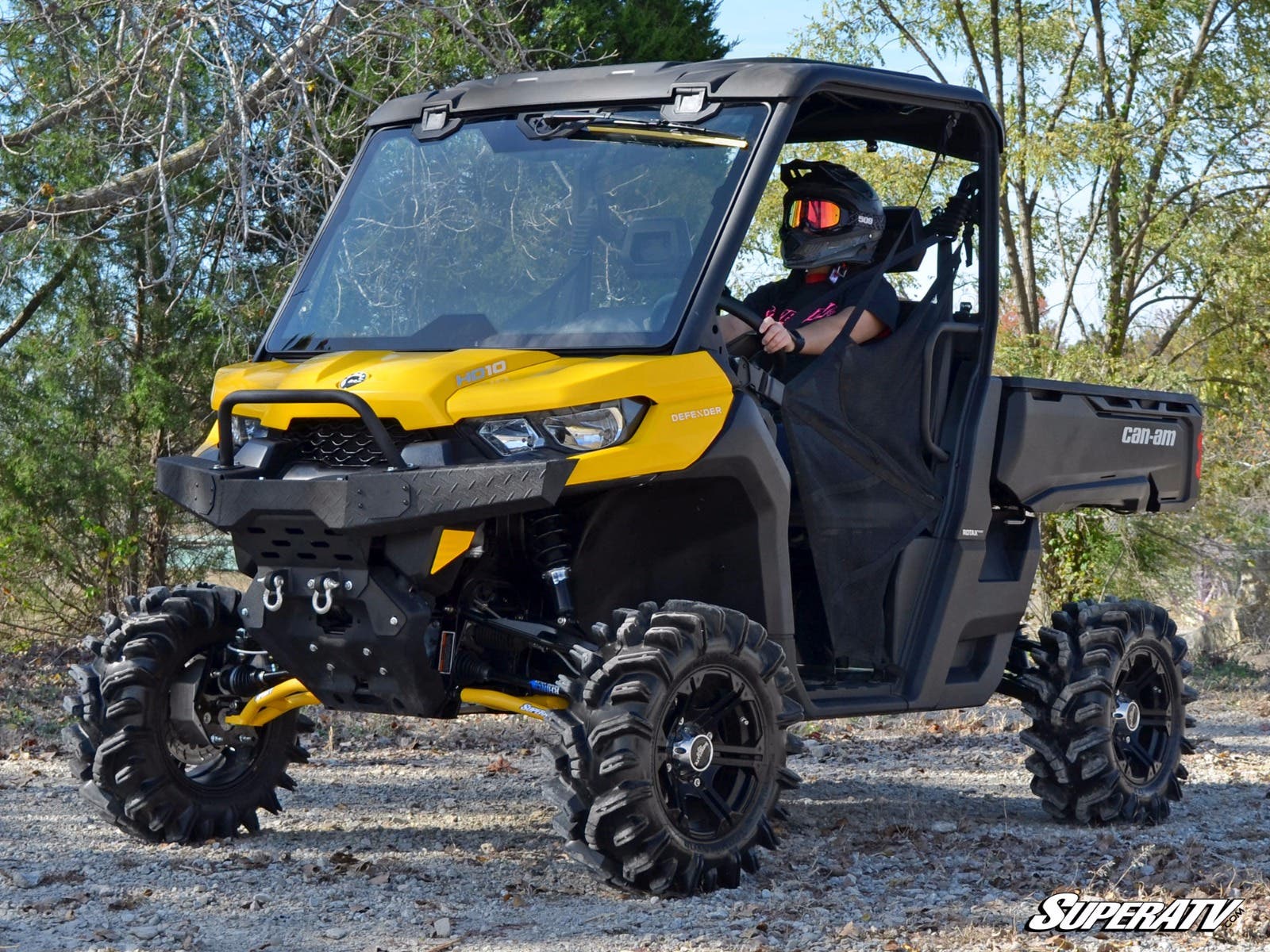 A person in a helmet and sunglasses drives a yellow and black Can-Am Defender equipped with the Can-Am Defender 4 Portal Gear Lift on a dirt path. The SuperATV logo appears in the lower right corner amid trees and grass.